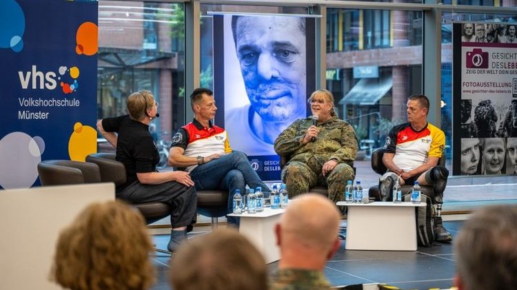 Das Bild wurde aus dem Publikum fotografiert. Es zeigt die Hinterköpfe des Publikums in Unschärfe und eine Talkrunde der VHS-Direktorin Esther Joy Dohmen mit drei „Gesichtern des Lebens“ zur Ausstellungseröffnung auf einer Bühne - v.l.n.r. Esther Joy Dohmen, Thomas Kroner, Claudia Schalling und Armin Witczak. Im Hintergrund des Fotos ist ein Roll-Up der VHS Münster sowie eins der „Gesichter-des-Lebens-Ausstellung“ zu erkennen und ein übergroßes Portrait-Foto von dem markanten Gesicht von Armin Witczak, das durch eine Sepsis gezeichnet ist.