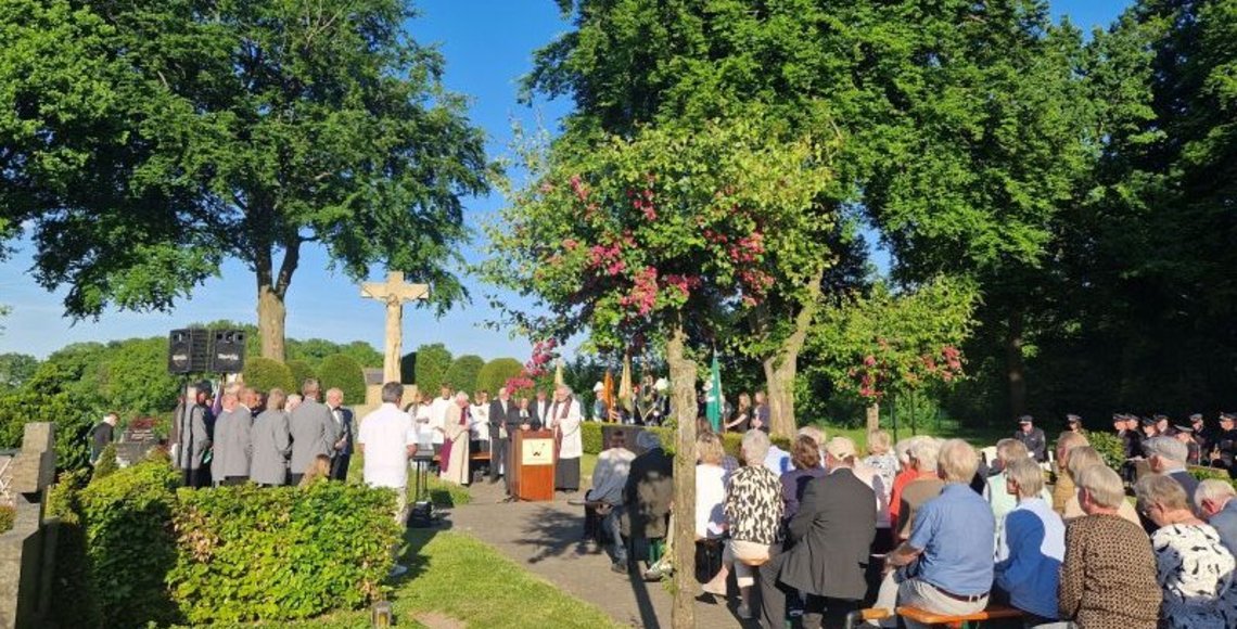 Auf dem Foto ist die Gedenkveranstaltung zum Kriegsende vor 80 Jahren auf dem Friedhof in Wettringen zu sehen. Im Vordergrund sieht man die Besucher auf einigen Bänken, links den Männergesangsverein, in der Mitte hinten das Rednerpult mit einem Wappen der Gemeinde, das Hochkreuz sowie rechts im Hintergrund den Musikzug der Freiwilligen Feuerwehr