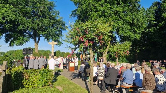 Auf dem Foto ist die Gedenkveranstaltung zum Kriegsende vor 80 Jahren auf dem Friedhof in Wettringen zu sehen. Im Vordergrund sieht man die Besucher auf einigen Bänken, links den Männergesangsverein, in der Mitte hinten das Rednerpult mit einem Wappen der Gemeinde, das Hochkreuz sowie rechts im Hintergrund den Musikzug der Freiwilligen Feuerwehr