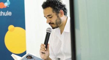 : Im Vordergrund ist der Autor, Dominik Wullers, mit einem strahlend weißen Hemd und einem schwarzen Mikrofon in der Hand zu sehen. Er liest aus seinem geöffneten Buch „Ich bin Deutschland. Plädoyer für einen liberalen Patriotismus“. Im Hintergrund sind etwas unscharf Teile des bunten Logos der VHS Münster zu erkennen.