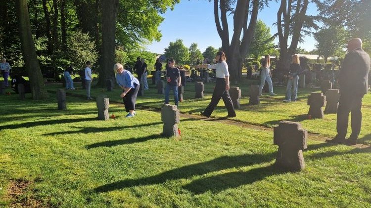 Das Foto zeigt die Realschülerinnen und -schüler mit einer Lehrerin und dem stellv. Bürgermeister Hartwig Guhle bei der Niederlegung von Gedenk-Kerzen an den 53 Kriegsgräber auf dem Friedhof. Die Abendsonne von Westen erzeugt lange Schatten der einzelnen, steinernen Grabkreuze.
