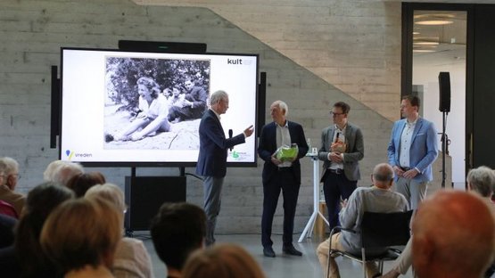 Über die Köpfe des Publikums hinweg ist links der Kreisdirektor des Kreises Borken, Dr. Ansgar Hörster zu sehen, der angesichts der doppelten Ausstellungseröffnung im kult Westmünsterland zwei Präsente an Paul Glaser (Initiator der Ausstellung „Die Tänzerin von Auschwitz“) und Jens Effkemann (Regionalgeschäftsführer des Volksbundes) verteilt. Daneben steht rechts der Bürgermeister der Stadt Vreden, Tom Tenostendarp. Im Hintergrund wird ein großes Foto der lachenden Tänzerin Roosje Glaser am Strand gezeigt. 